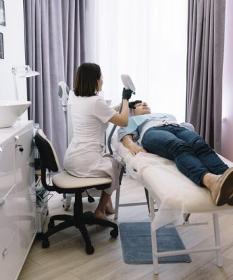 Professional cosmetician in uniform using YAG laser for photo epilation on female clients face lying on couch in modern beauty clinic